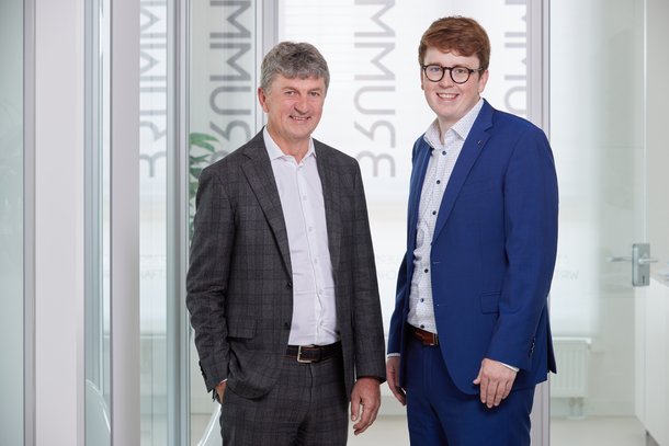 Zwei Männer stehen nebeneinander und lächeln in die Kamera. Sie tragen Anzüge; der ältere Mann einen dunklen, der jüngere einen blauen. Im Hintergrund sind Glaswände mit Schrift.