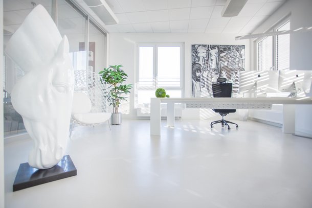 Eine weiße Pferdeskulptur steht in einem modernen, hellen Büro mit einem Schreibtisch und Bürostuhl vor einem großen Fenster, einem Kunstwerk an der Wand und einer Zimmerpflanze.