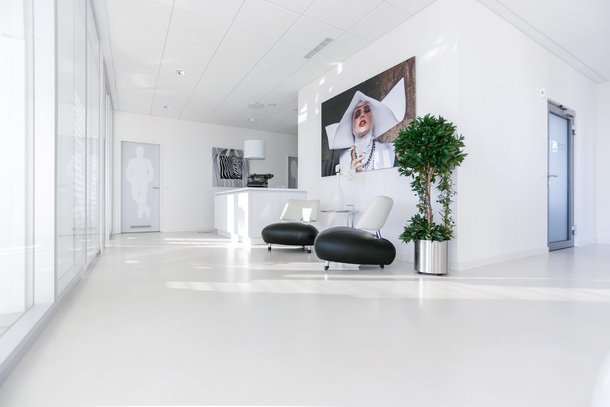 Zwei moderne schwarze Stühle stehen in einem hellen, minimalistisch eingerichteten Raum. Eine große Kunstfotografie hängt an der Wand, neben einer grünen Topfpflanze. Links ist eine Glaswand sichtbar.